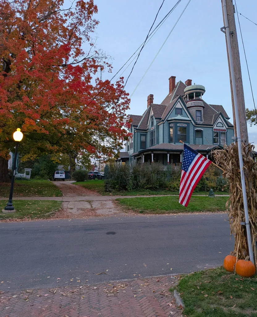 A blue Victorian house with a turret and wraparound porch behind a red maple near Bradley University