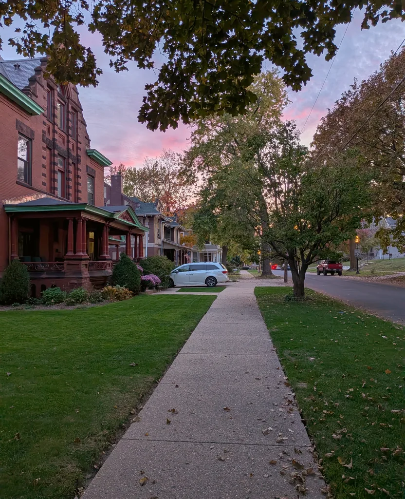 A sidewalk past a red brick Victorian with green trim under a pink sunset sky in Peoria's West Bluff