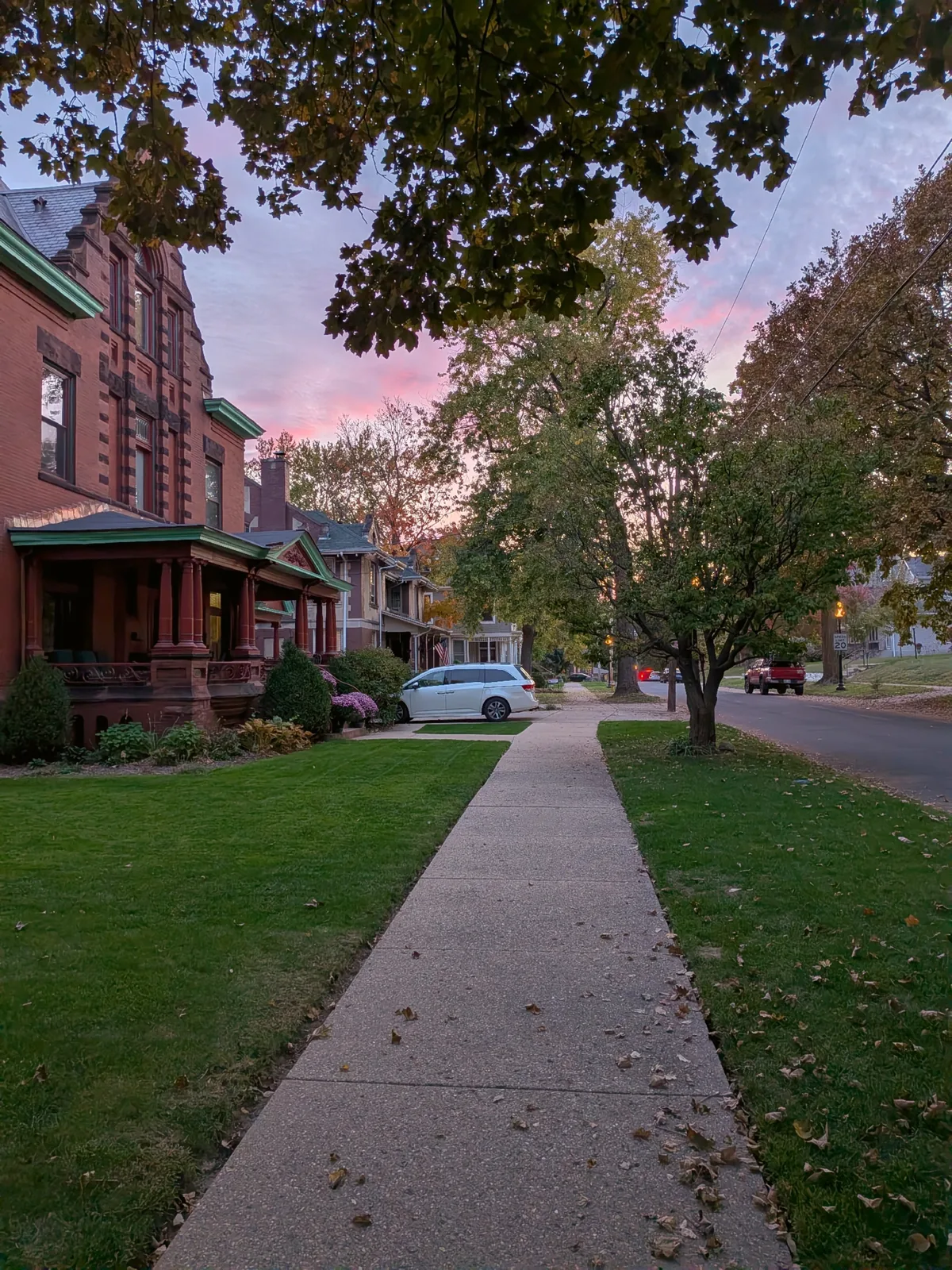 A row of brick Victorians with front porches along a sidewalk at pink sunset