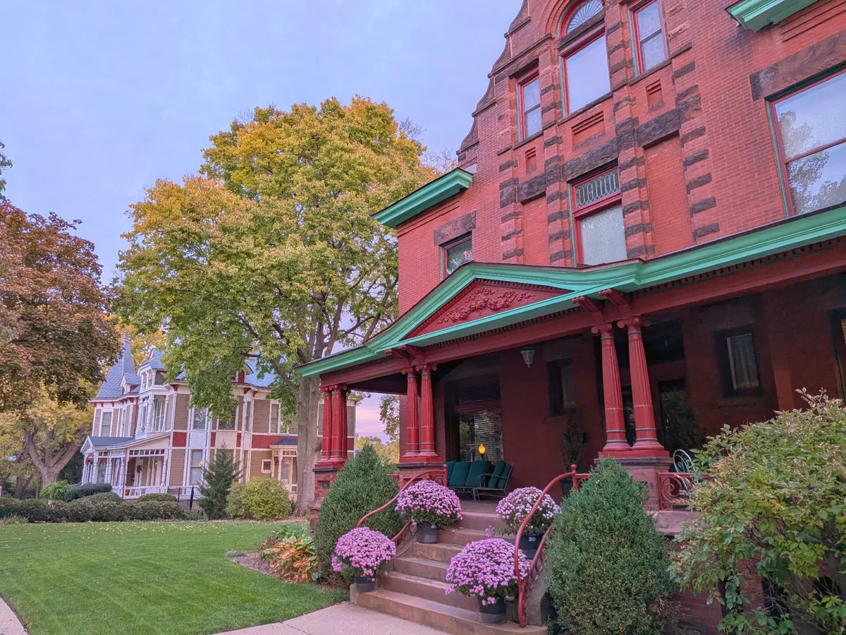 A red brick Romanesque house with green trim and pink mums on the steps, beside a painted Victorian